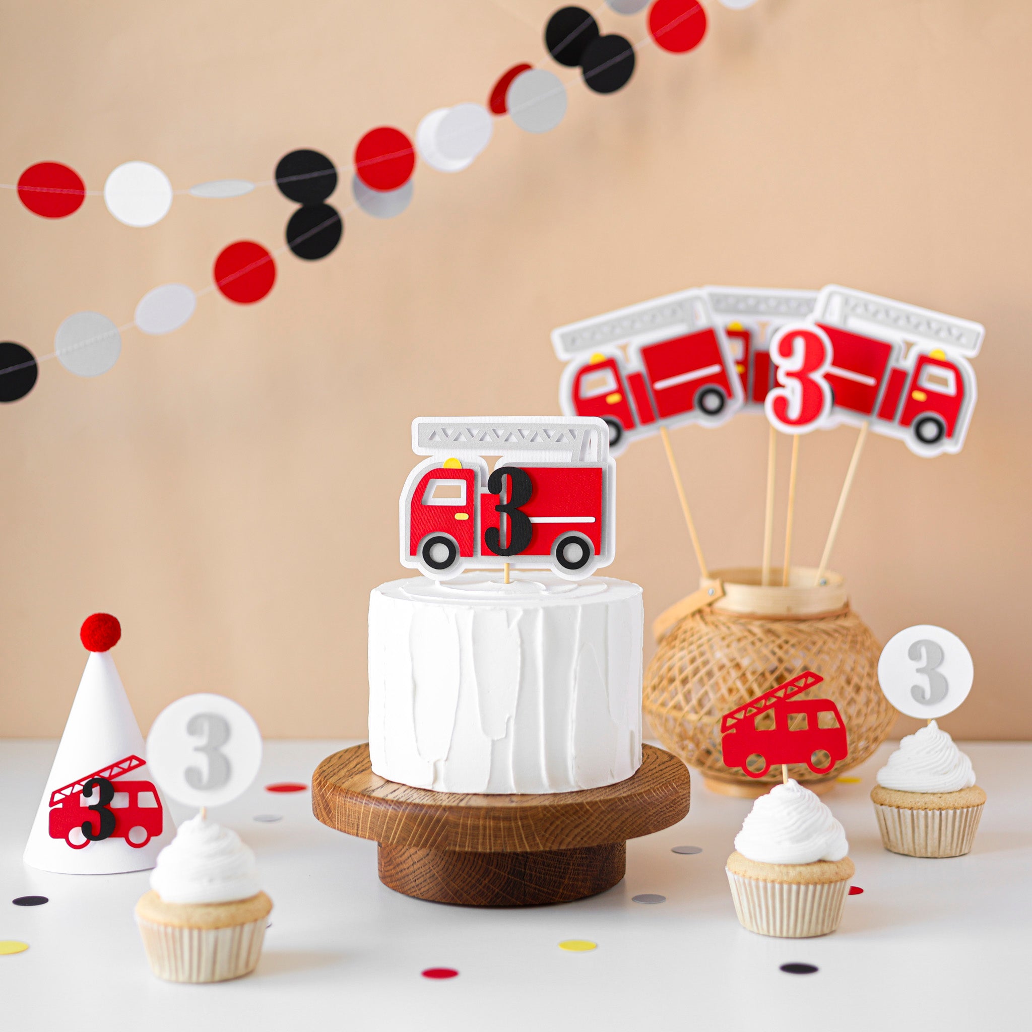 A festive firefighter-themed birthday setup featuring a white cake with a fire truck topper, coordinated cupcakes with number "3" and fire engine picks, a matching party hat, fire truck centerpieces, and a red, white, and black circle paper garland.