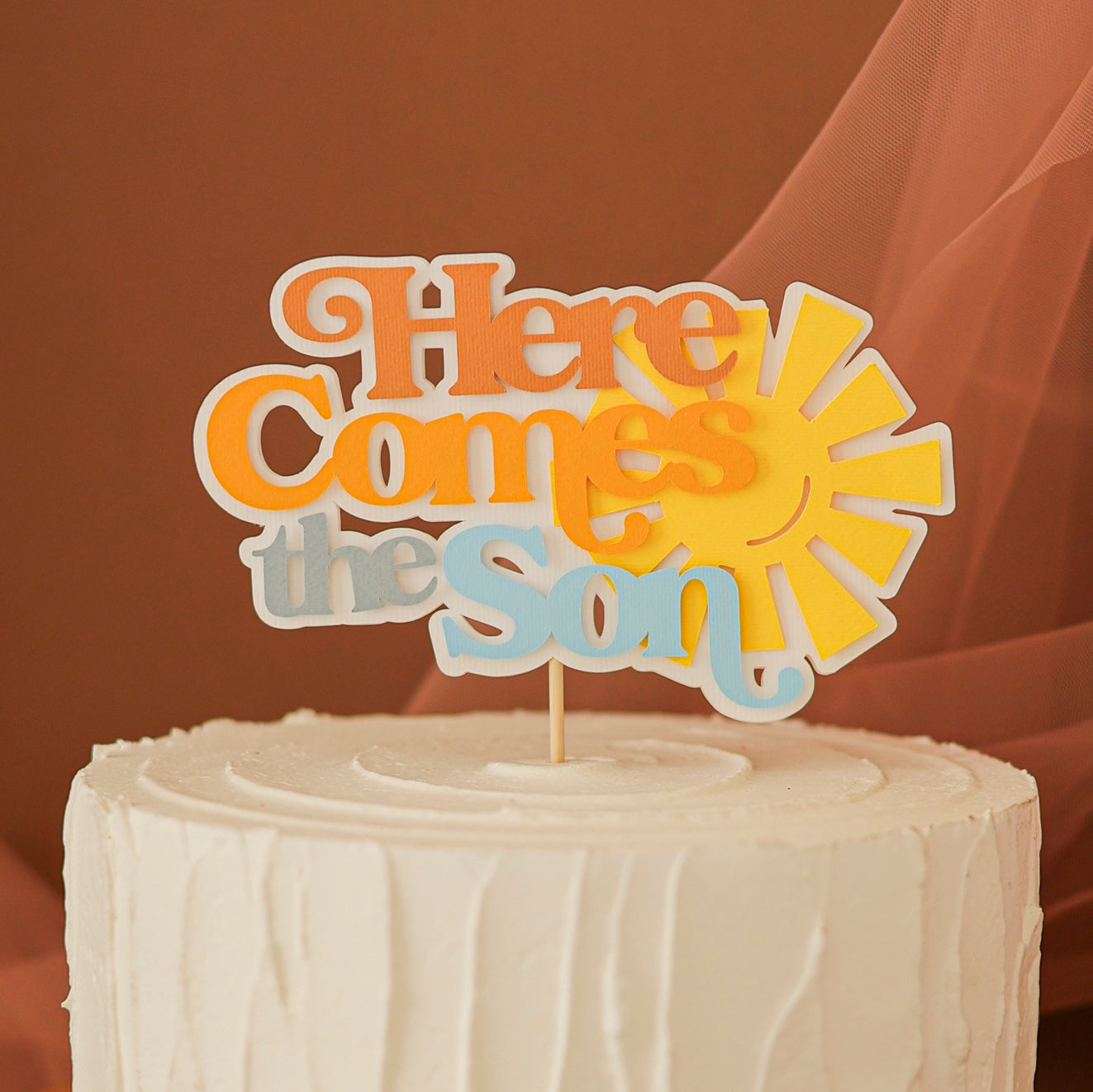 "Here Comes The Son" cake topper in orange, yellow, and light blue letters with a yellow sun icon, placed on top of a white frosted cake, suitable for a baby shower or birthday