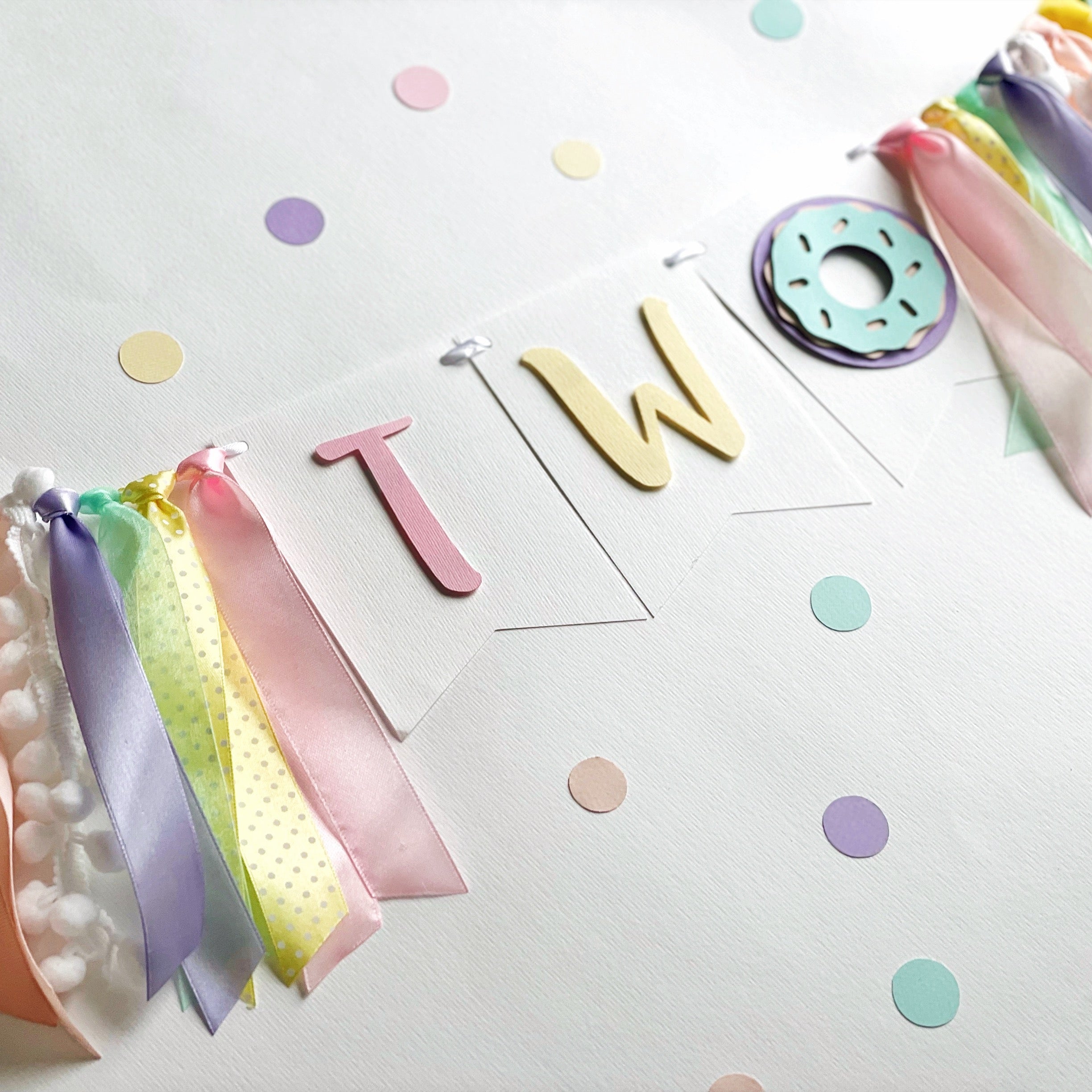 pastel birthday party decoration set featuring a 'TWO' banner with ribbon tassels, a donut decoration, and scattered pastel confetti 