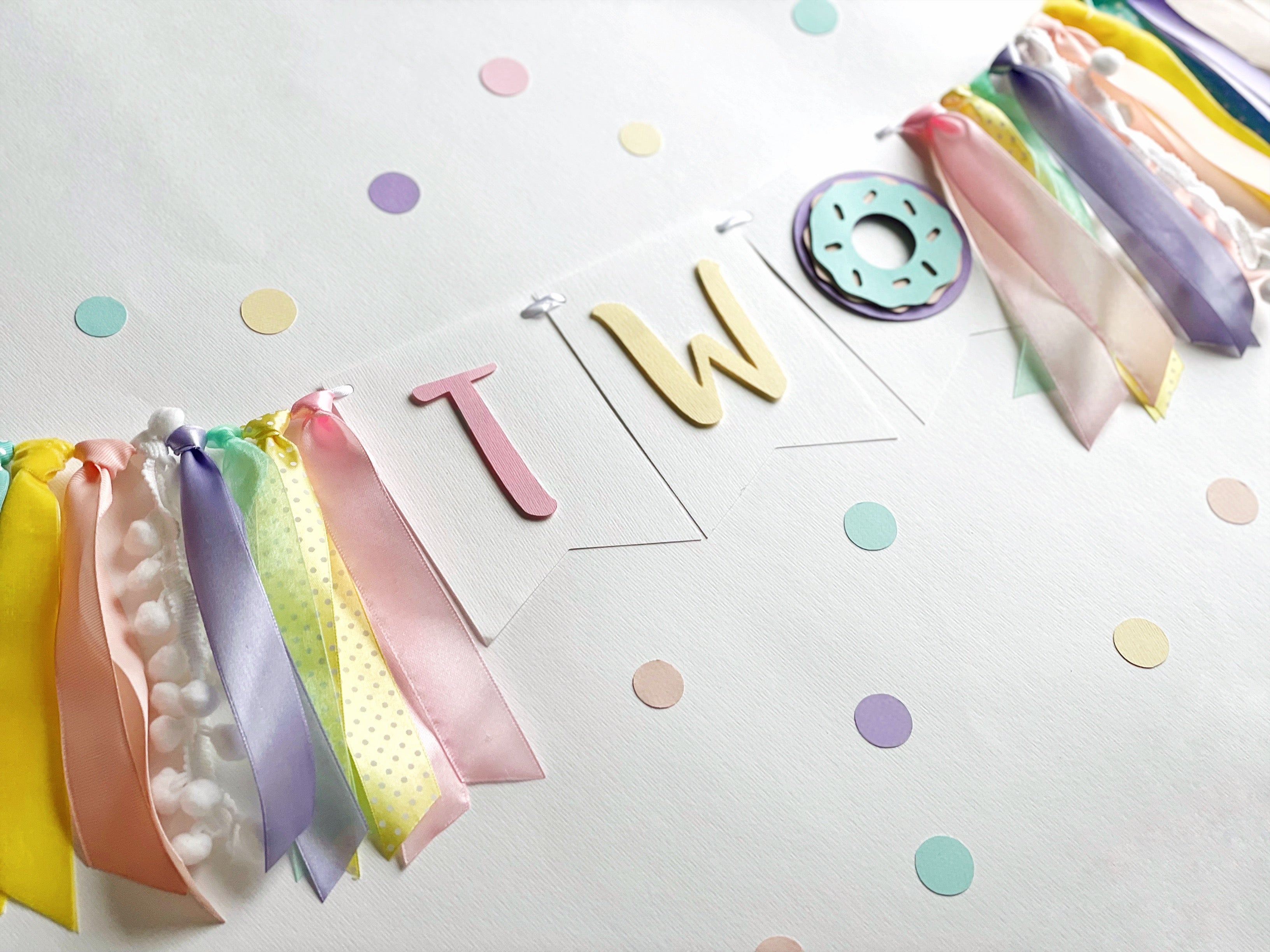 pastel birthday party decoration set featuring a 'TWO' banner with ribbon tassels, a donut decoration, and scattered pastel confetti 