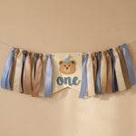 Handmade bear highchair banner for boys 1st birthday featuring blue, beige, and brown ribbons with a bear face and "one" cutout.