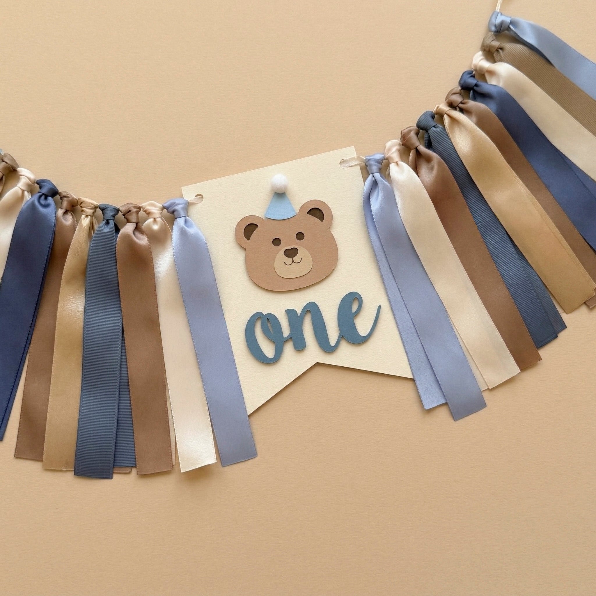 Handmade bear highchair banner for boys 1st birthday featuring blue, beige, and brown ribbons with a bear face and "one" cutout.