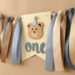Handmade bear highchair banner for boys 1st birthday featuring blue, beige, and brown ribbons with a bear face and "one" cutout.