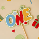 Colorful 'ONE' letter cake topper on a stick with decorative elements on a beige background