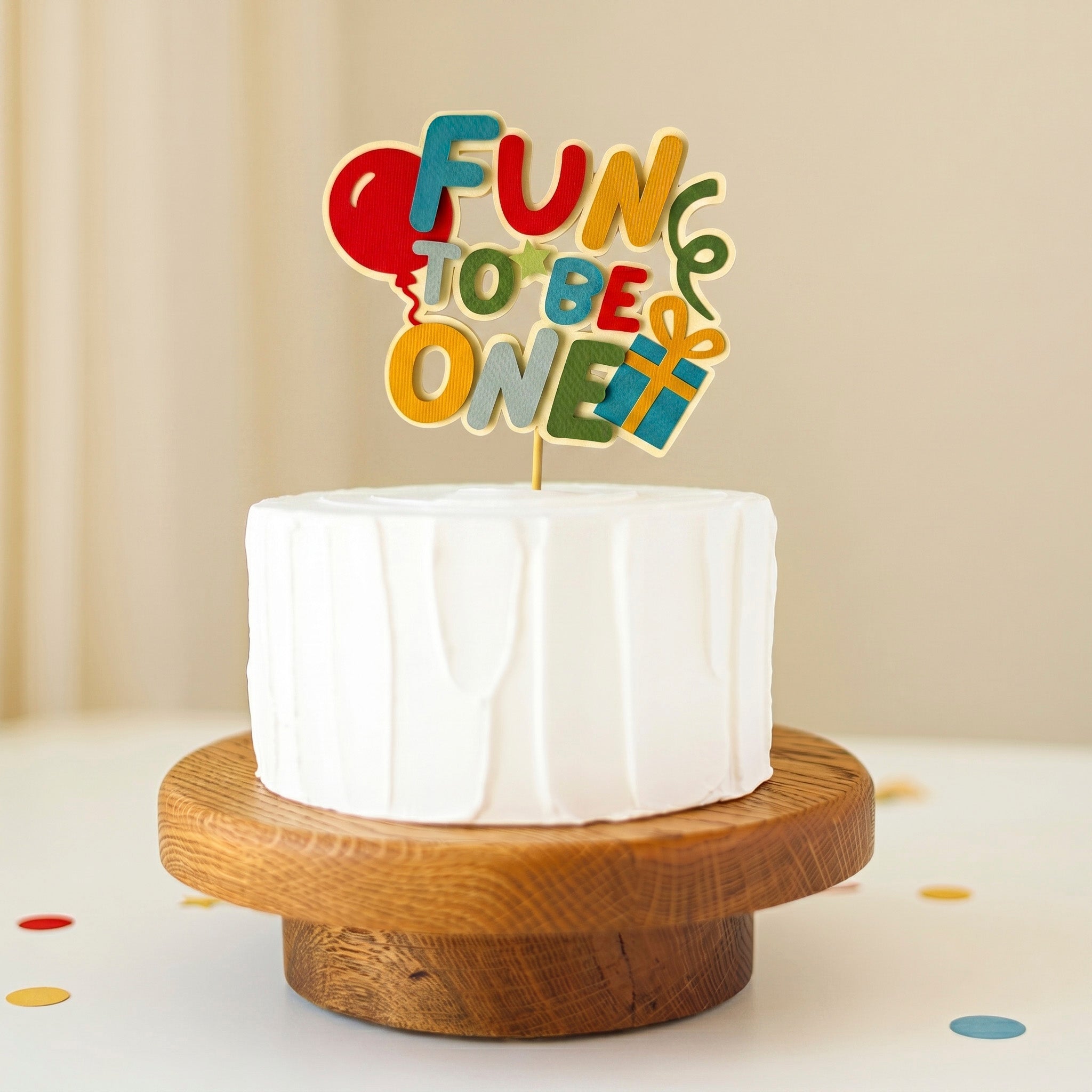 White cake on a wooden stand with a colorful 'Fun to be One' cake topper against a beige background.