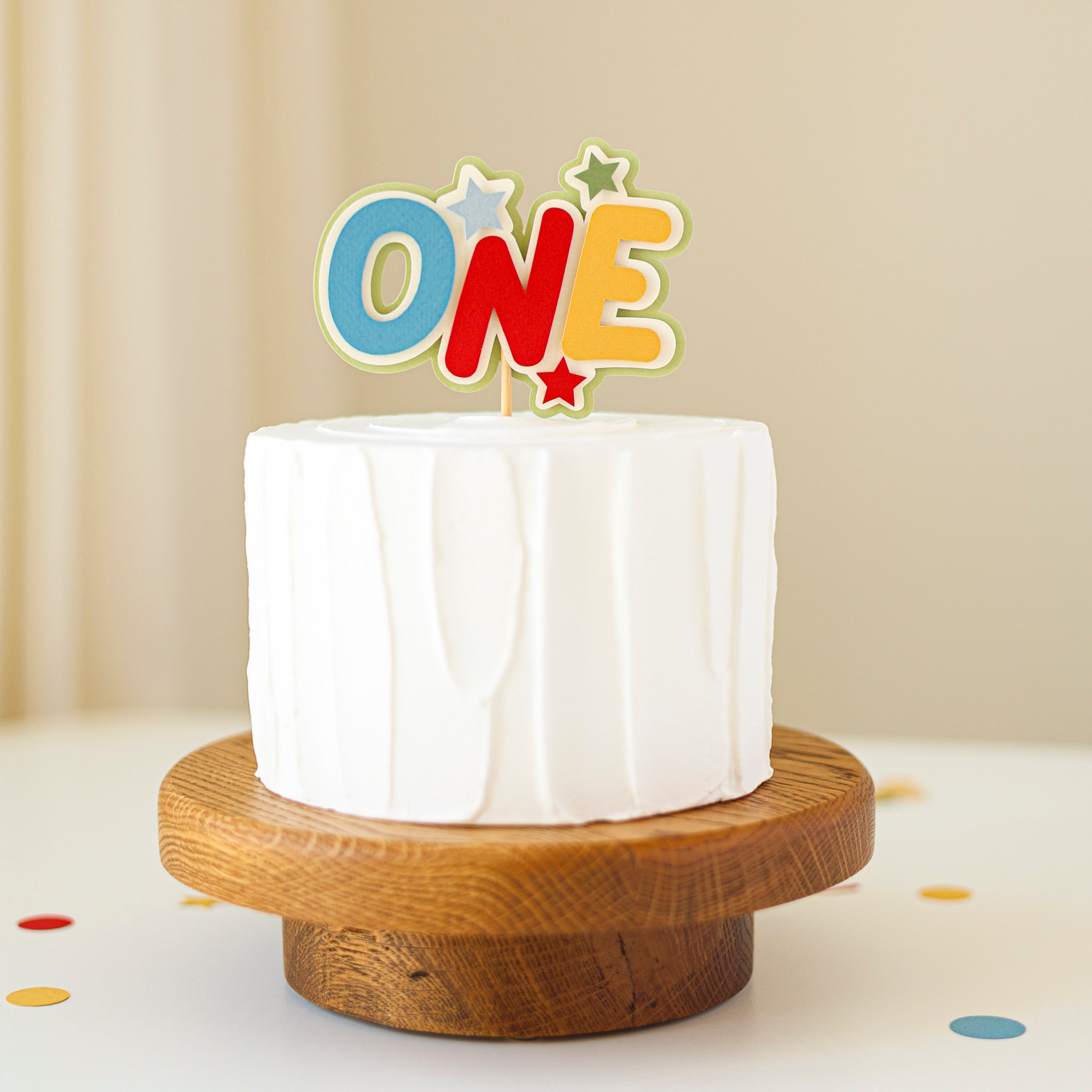 Small white cake on a wooden stand with a colorful 'ONE' topper against a beige background