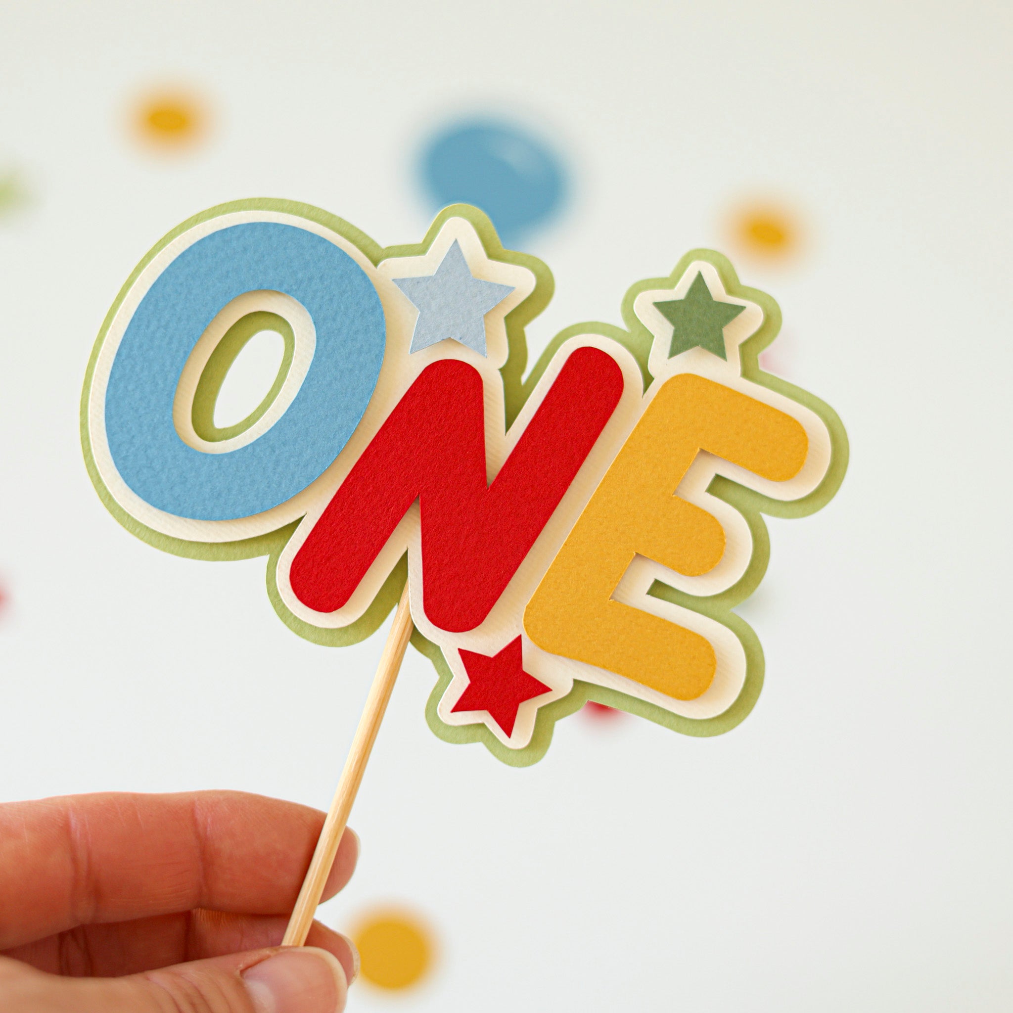 Colorful 'ONE' cake topper on a stick held by a hand against a light background.