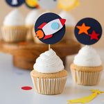 Handcrafted "First Trip Around the Sun" cupcake toppers with navy blue circles, white number ones, a yellow sun, a grey moon, and a red rocket ship on a white background.