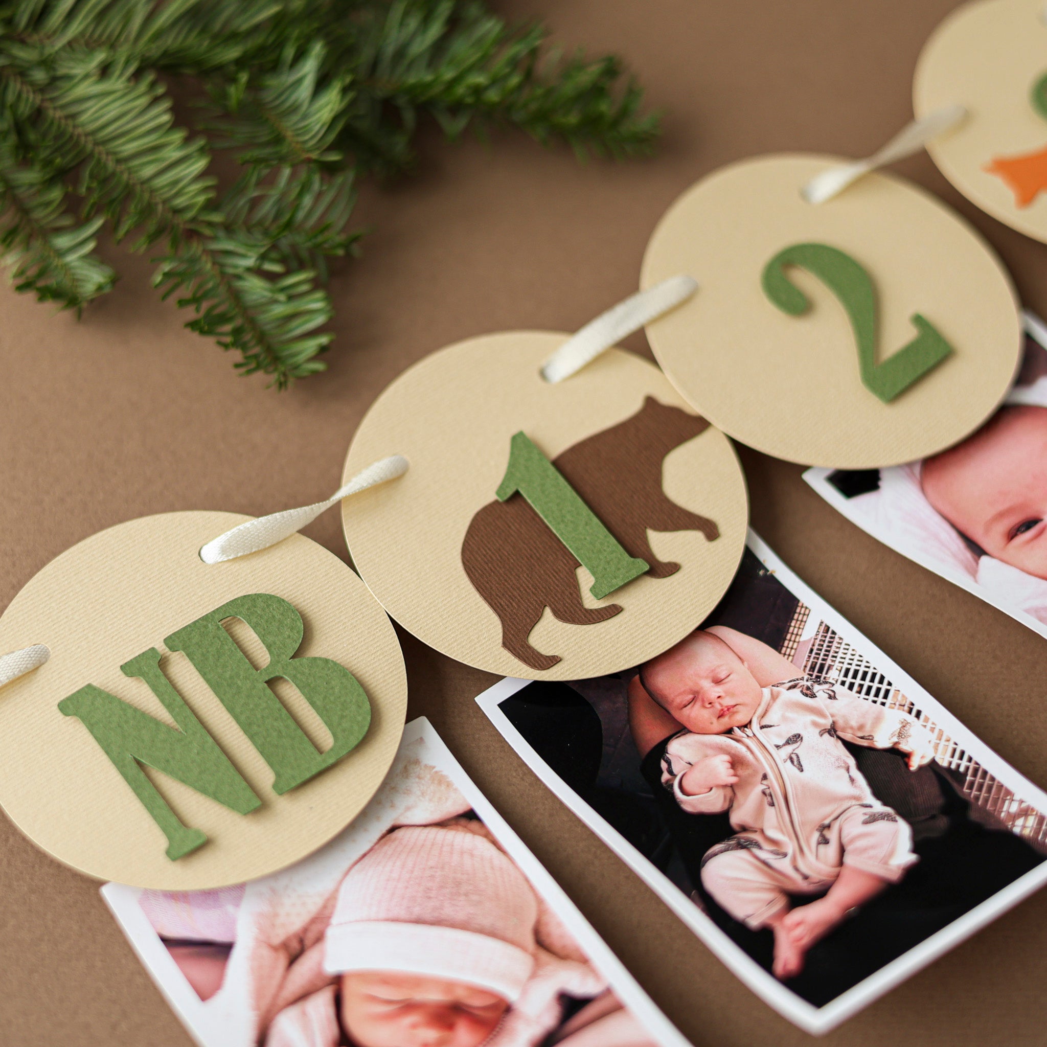 Close-up of woodland birthday photo garland featuring a bear silhouette and "1 Month" card in earthy green and brown tones.