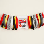 Firetruck 1st birthday highchair banner with red, yellow, and black ribbons featuring a 3D firefighter truck and "one" sign for a boy's cake smash.