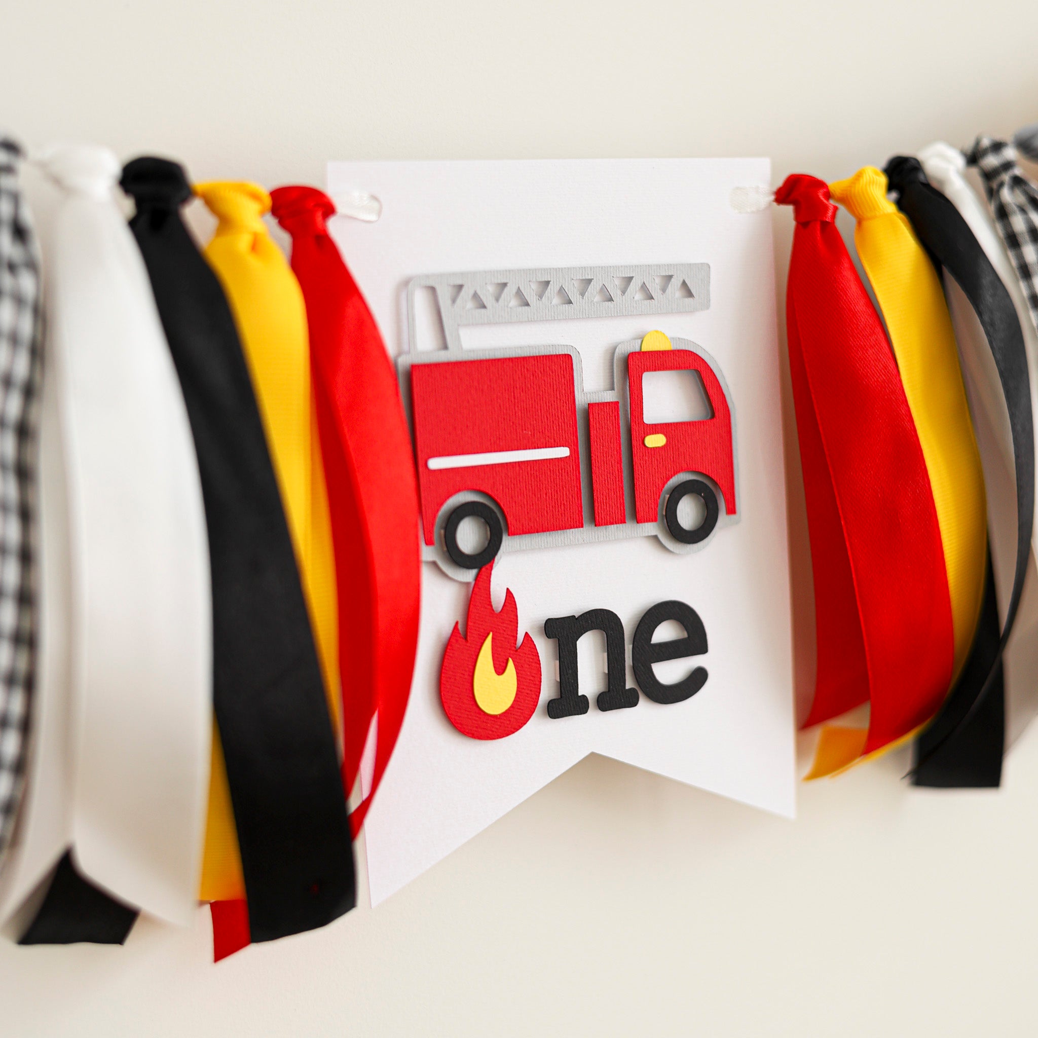 Firetruck 1st birthday highchair banner with red, yellow, and black ribbons featuring a 3D firefighter truck and "one" sign for a boy's cake smash.