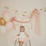 Child in a high chair with 'Happy Birthday' banner and balloons, celebrating a special occasion.