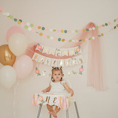 Child in a high chair with 'Happy Birthday' banner and balloons, celebrating a special occasion.