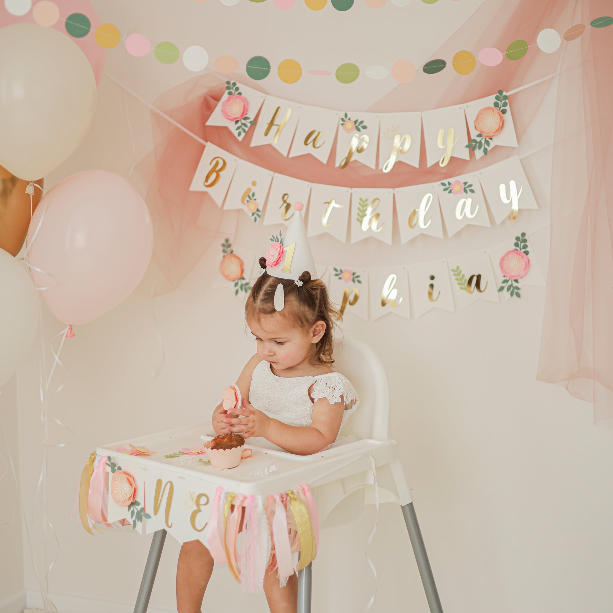 Child in a high chair with 'Happy Birthday' banner and balloons, celebrating a first birthday.