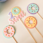 Four pastel donut-shaped cake toppers on wooden sticks arranged on a light cream background, with a pink and white script word sign reading "Sweet" between two of the toppers.