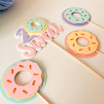 A set of pastel donut-shaped party toppers on wooden sticks, featuring layered pink, yellow, and mint green donut designs with the word 'Sweet' in layered script letters.
