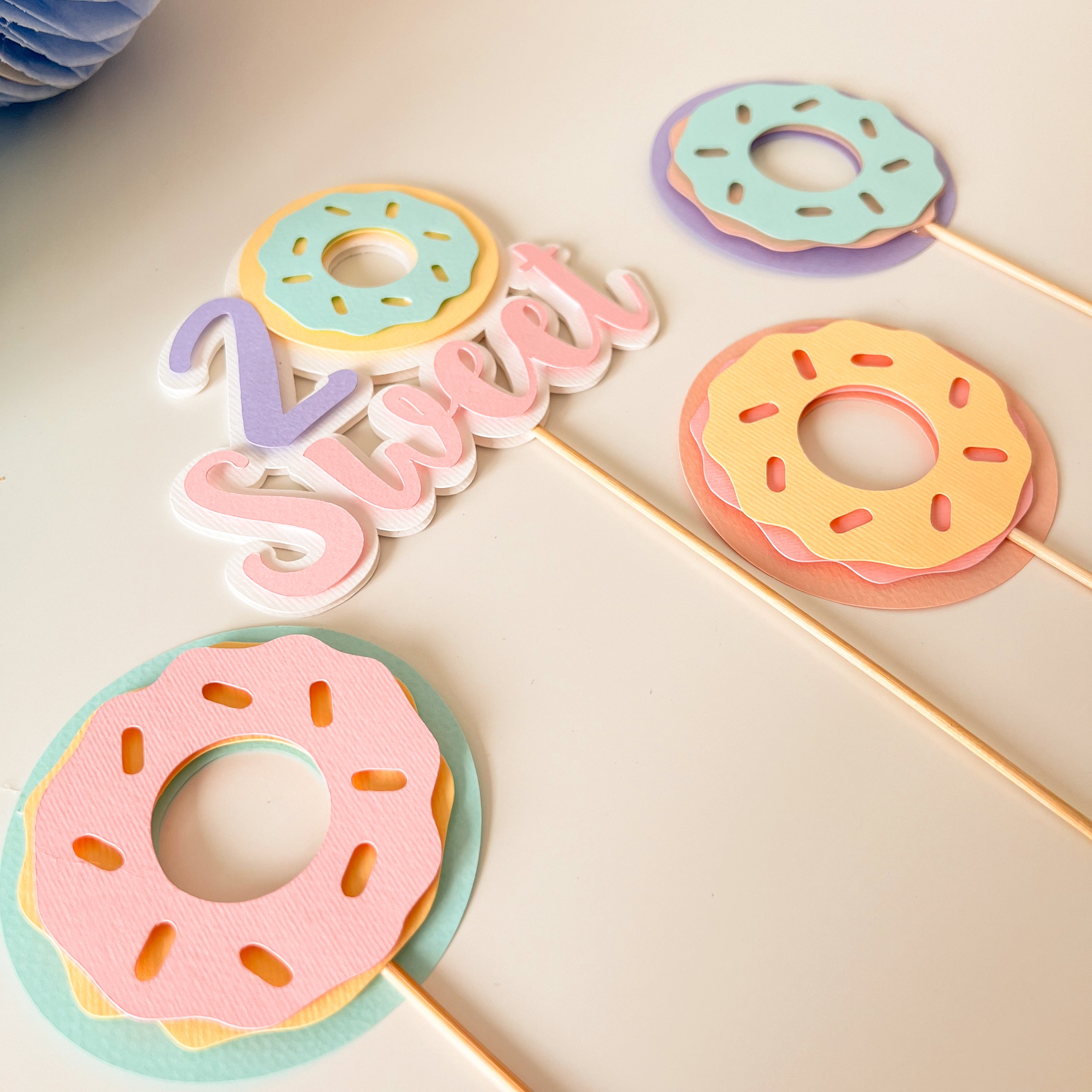 A set of pastel donut-shaped party toppers on wooden sticks, featuring layered pink, yellow, and mint green donut designs with the word 'Sweet' in layered script letters.