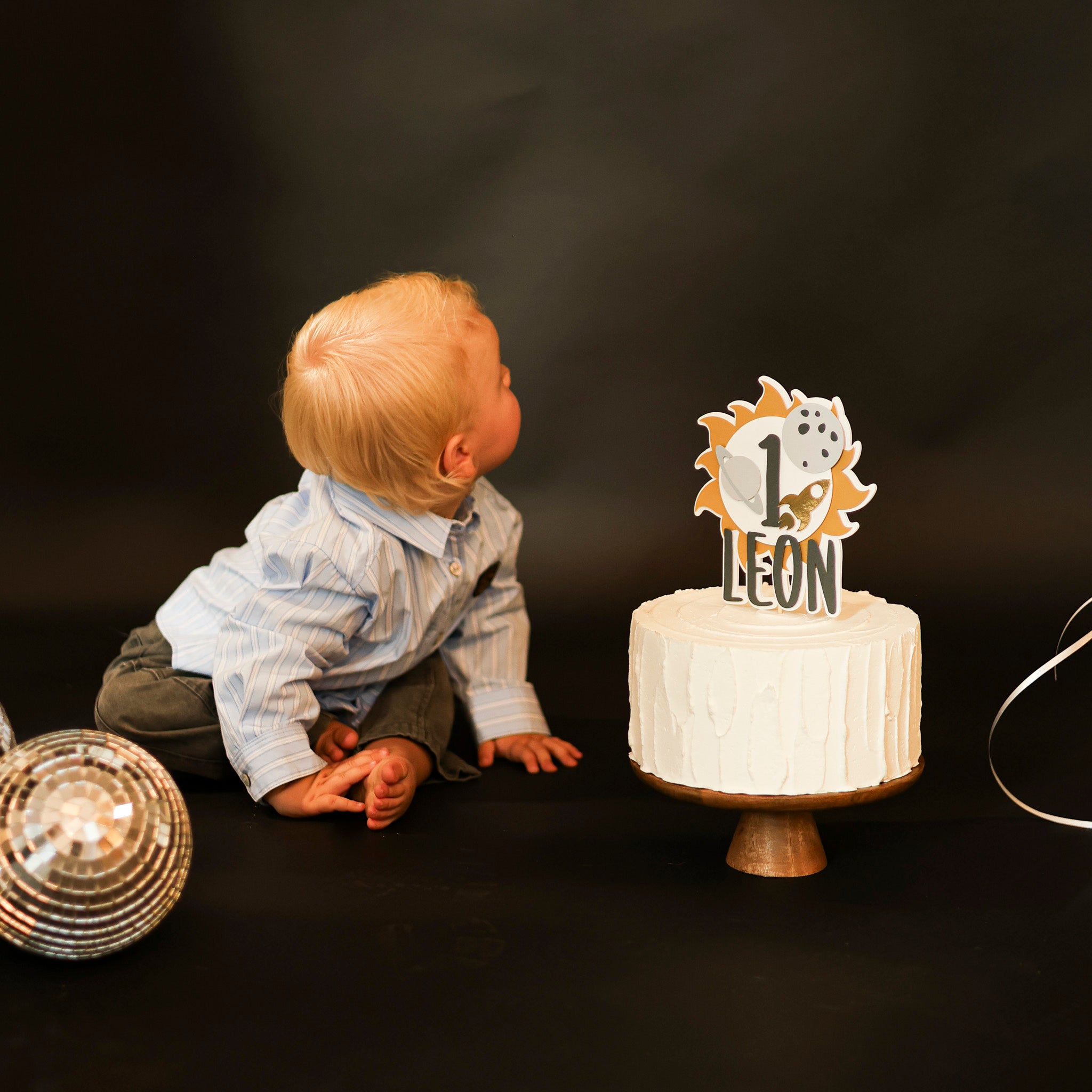 Personalized "First Trip Around the Sun" cake topper for a 1st birthday, featuring the number '1', a golden sun, grey planet, moon, and gold rocket ship, placed on a white frosted cake 