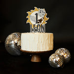 Personalized "First Trip Around the Sun" cake topper for a 1st birthday, featuring the number '1', a golden sun, grey planet, moon, and gold rocket ship, with the baby's name in dark grey letters, placed on a white frosted cake on a wooden stand