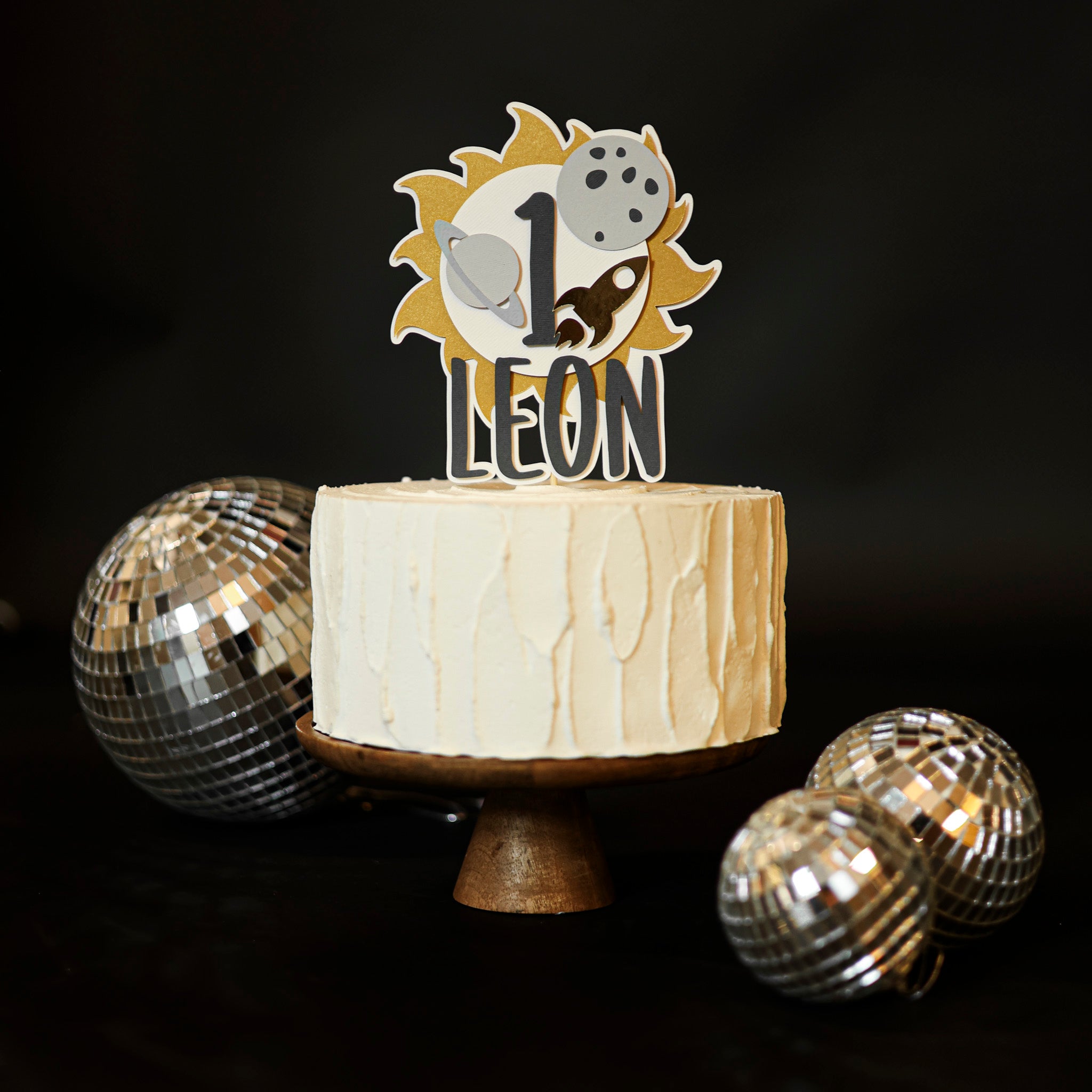 Personalized "First Trip Around the Sun" cake topper for a 1st birthday, featuring the number '1', a golden sun, grey planet, moon, and gold rocket ship, with the baby's name in dark grey letters, placed on a white frosted cake on a wooden stand