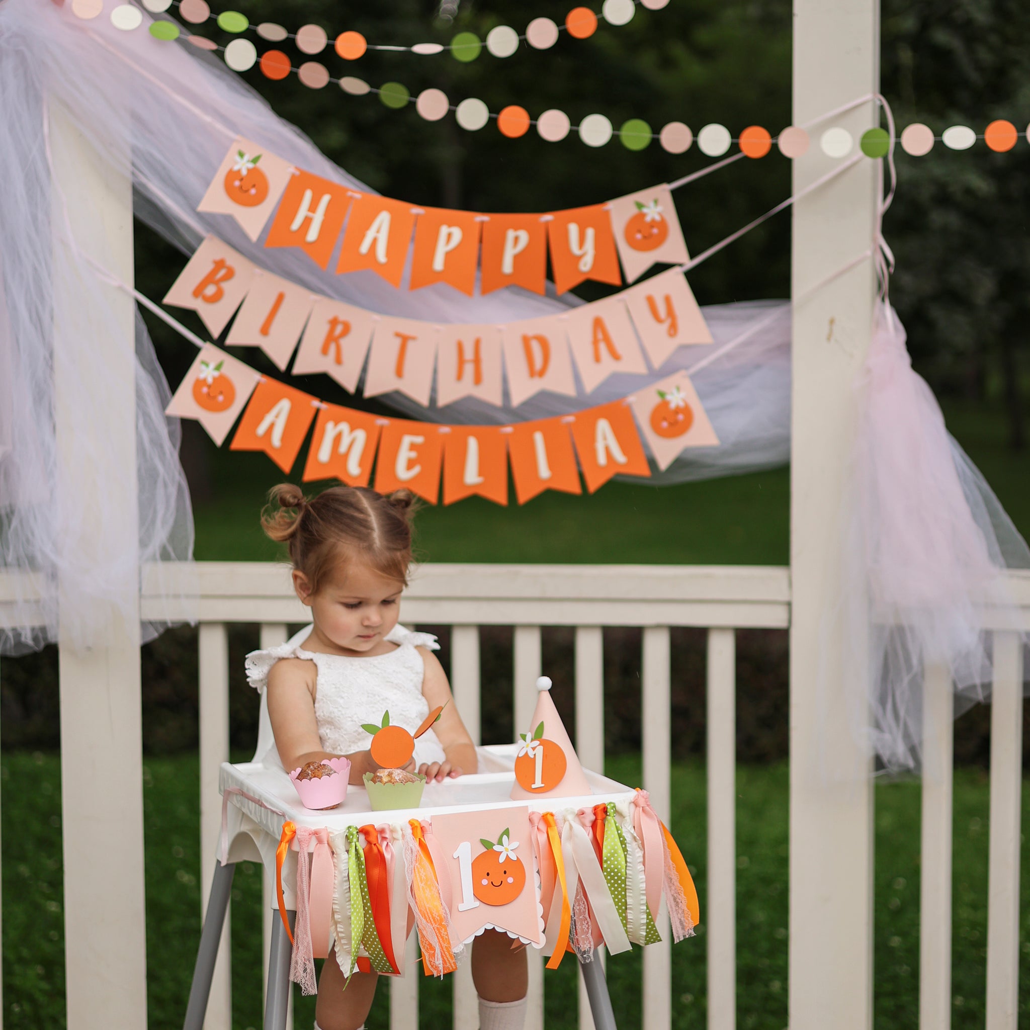 "Happy Birthday Amelia" banner in orange and pink with orange accents, hanging above a highchair decorated with matching orange and green ribbons and an orange-themed "1" banner. A toddler girl with a cupcake is sitting in the highchair and a party hat, at an outdoor "Little Cutie" or orange-themed first birthday party setup.