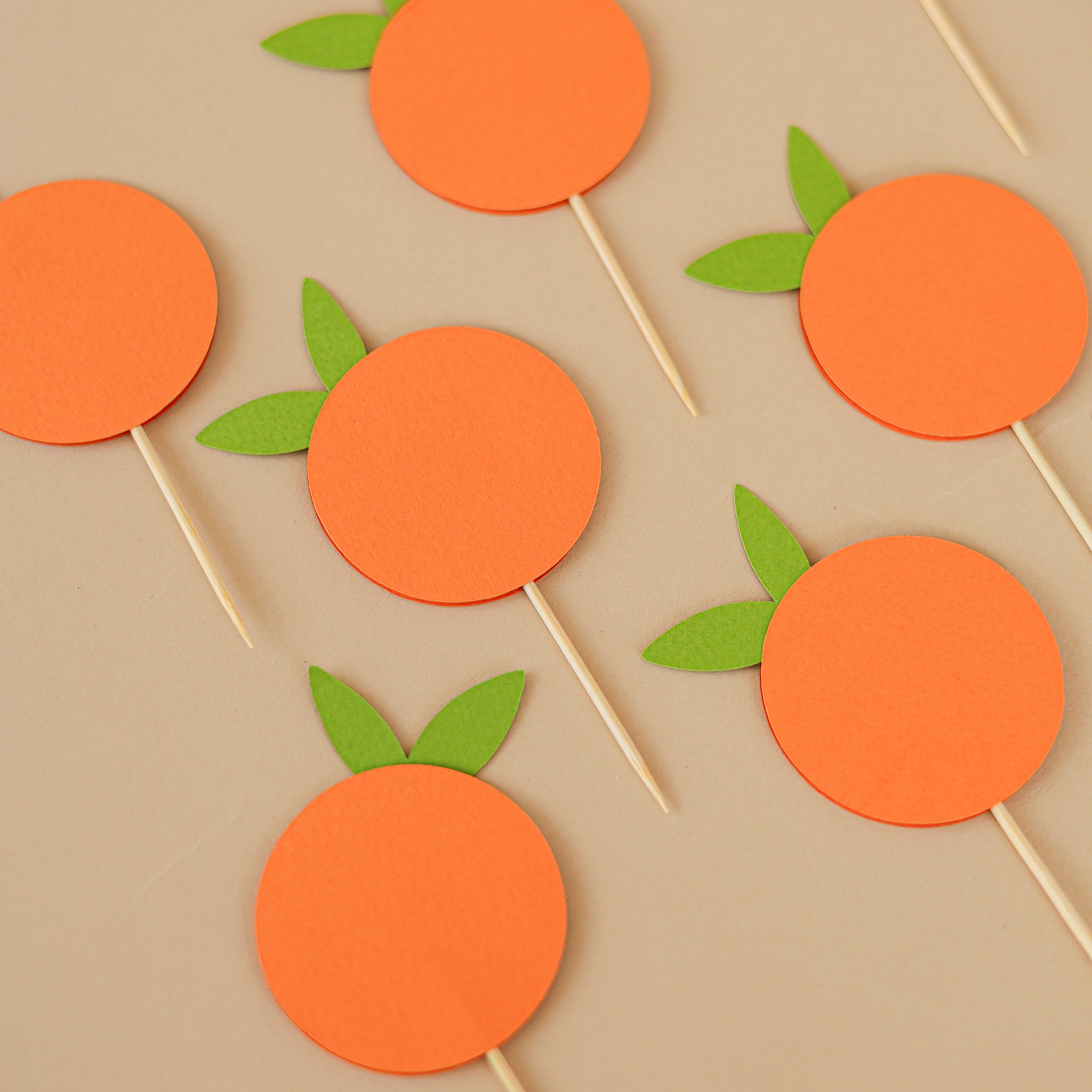 "Little Cutie" cupcake toppers, featuring bright orange circle cutouts with green leaves on wooden sticks, ideal for baby shower or 1st birthday party decorations