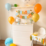 A styled children's birthday party setup featuring a white dresser decorated with a colorful 'Happy Birthday' banner, balloons in blue, orange, and yellow, honeycomb paper decorations on the floor, and a white high chair with a matching banner.