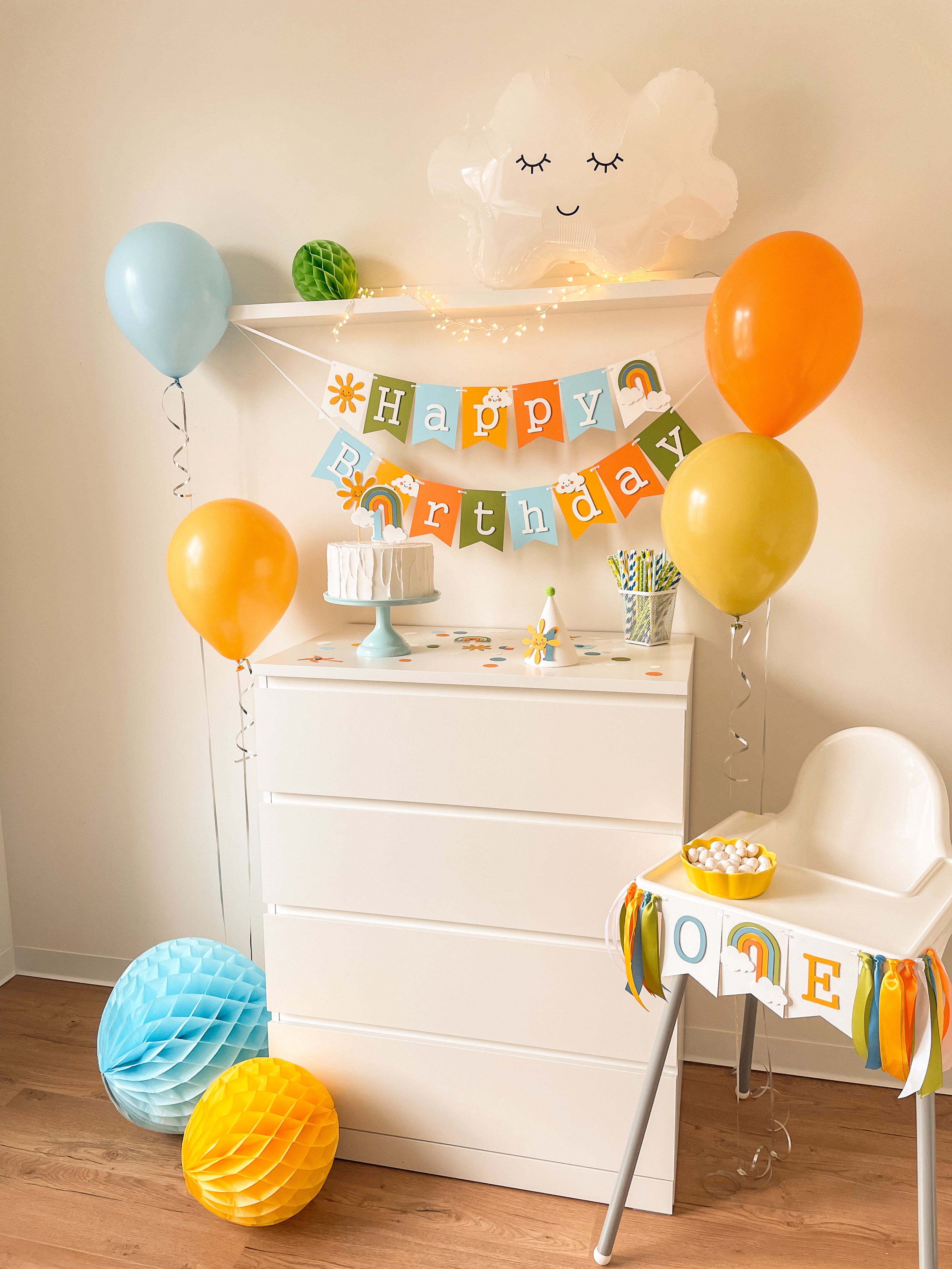 A styled children's birthday party setup featuring a white dresser decorated with a colorful 'Happy Birthday' banner, balloons in blue, orange, and yellow, honeycomb paper decorations on the floor, and a white high chair with a matching banner.