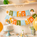Handmade first birthday party hat with smiling yellow sun, blue number 1, and green pom pom on light blue cake stand with pastel garland background.
