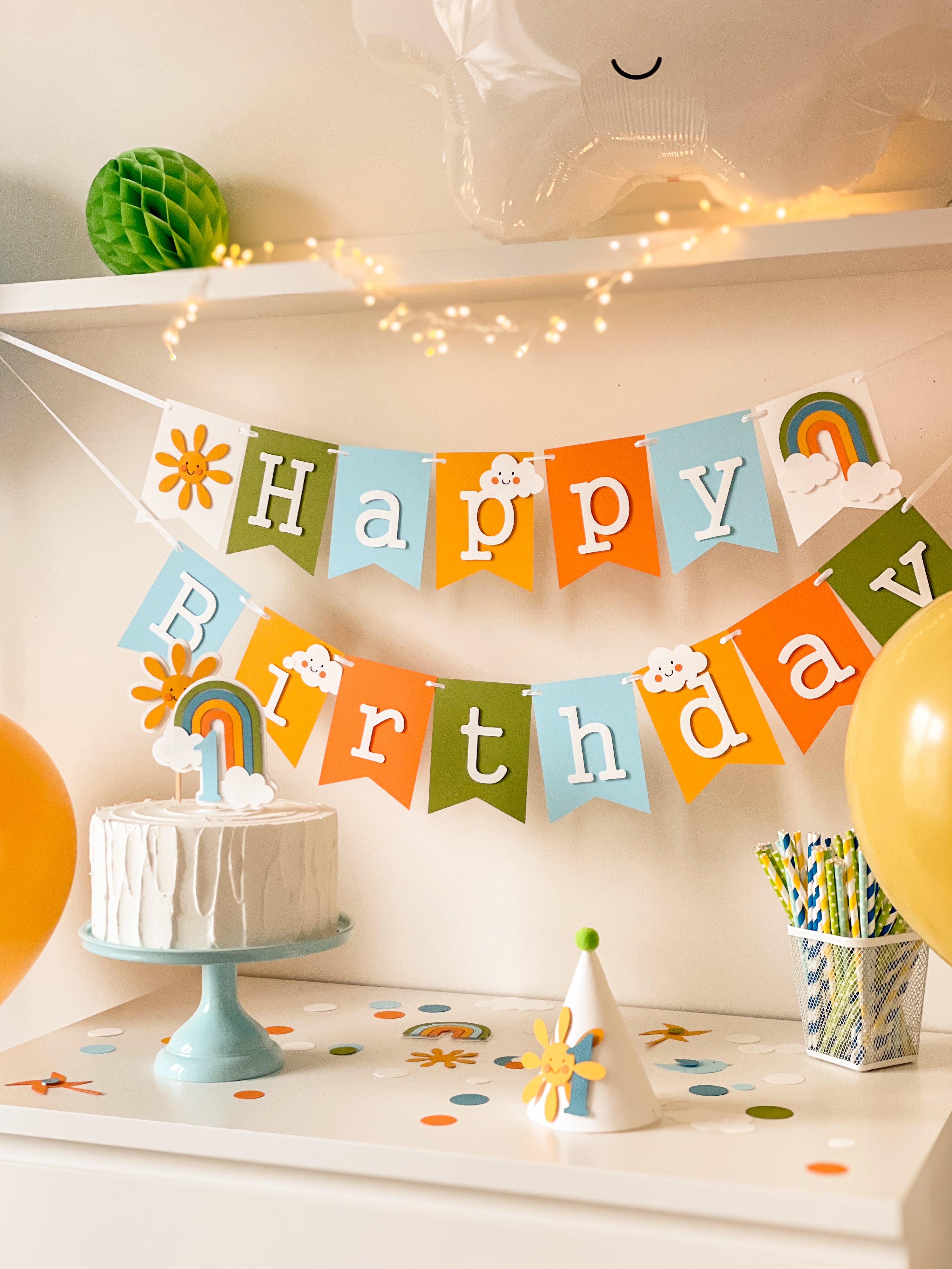 Handmade first birthday party hat with smiling yellow sun, blue number 1, and green pom pom on light blue cake stand with pastel garland background.