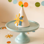 Handmade first birthday party hat with smiling yellow sun, blue number 1, and green pom pom on light blue cake stand with pastel garland background.