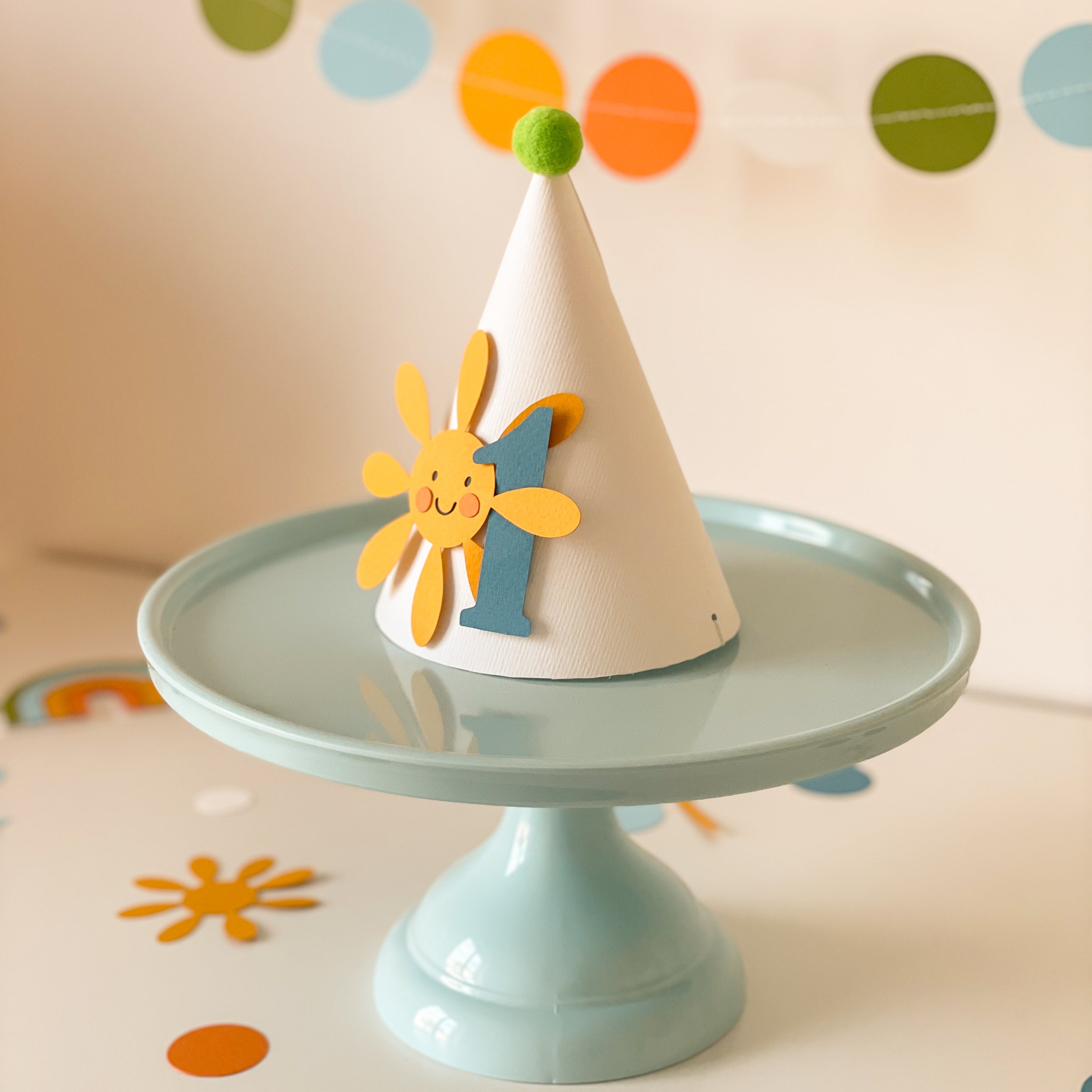 Handmade first birthday party hat with smiling yellow sun, blue number 1, and green pom pom on light blue cake stand with pastel garland background.