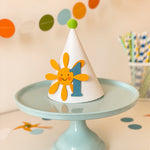 A white party hat with a smiling yellow sun and blue number 1 decoration, displayed on a light blue cake stand against a softly blurred party background with colorful balloons and straws.