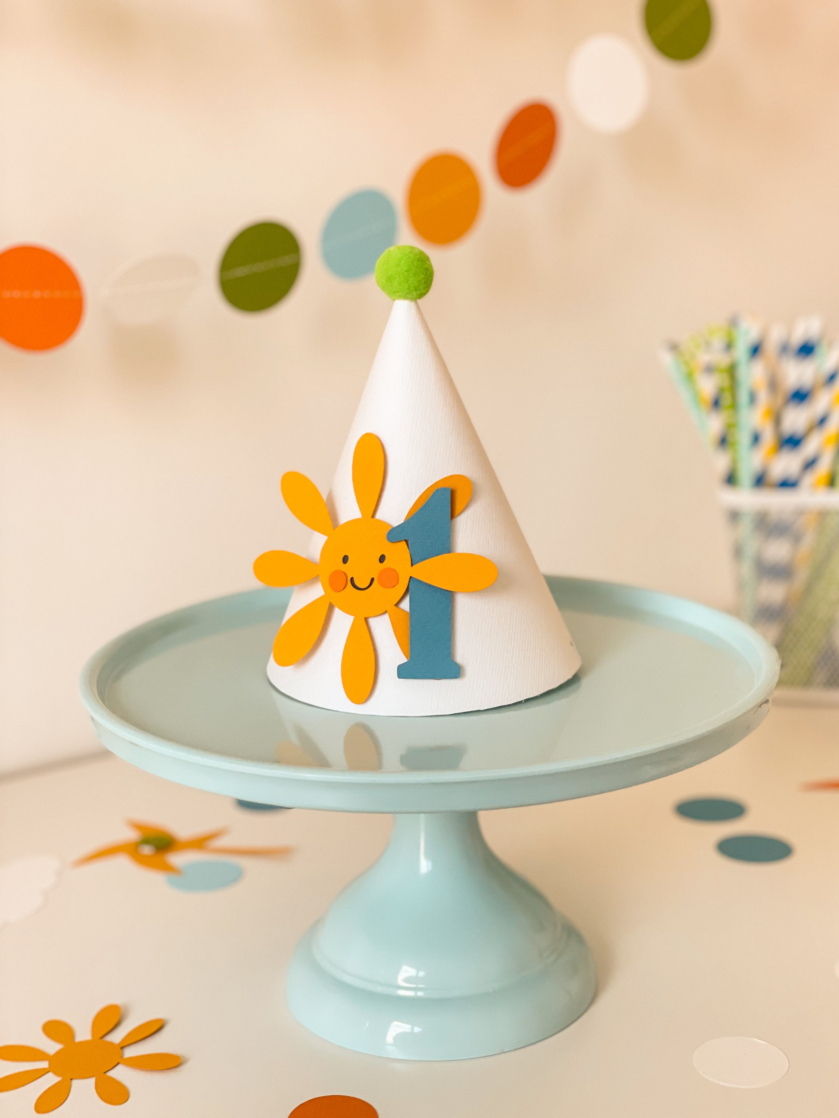 A white party hat with a smiling yellow sun and blue number 1 decoration, displayed on a light blue cake stand against a softly blurred party background with colorful balloons and straws.