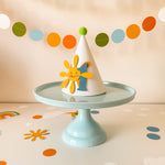 A white party hat with a smiling yellow sun and blue number 1 decoration, displayed on a light blue cake stand against a warm beige background with colorful confetti and a paper garland.