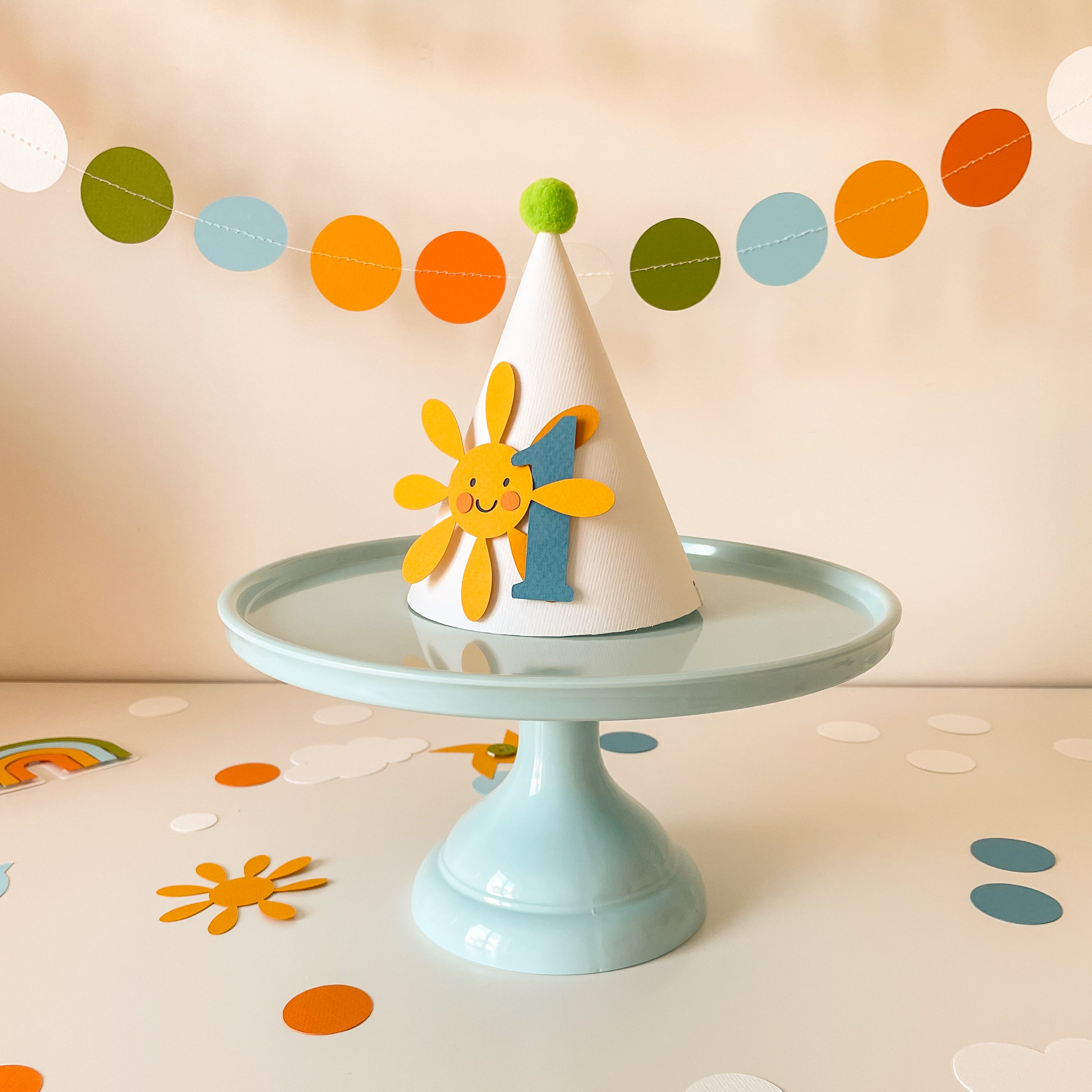 A white party hat with a smiling yellow sun and blue number 1 decoration, displayed on a light blue cake stand against a warm beige background with colorful confetti and a paper garland.