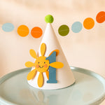 Handmade first birthday party hat with smiling yellow sun, blue number 1, and green pom pom on light blue cake stand with pastel garland background.