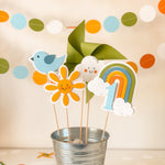 Colorful party decorations including a bird, flower, cloud, and rainbow in a metal bucket with a polka dot background.