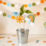 Decorative items including a number '1', rainbow, cloud, and sun in a metal bucket with colorful paper decorations in the background.