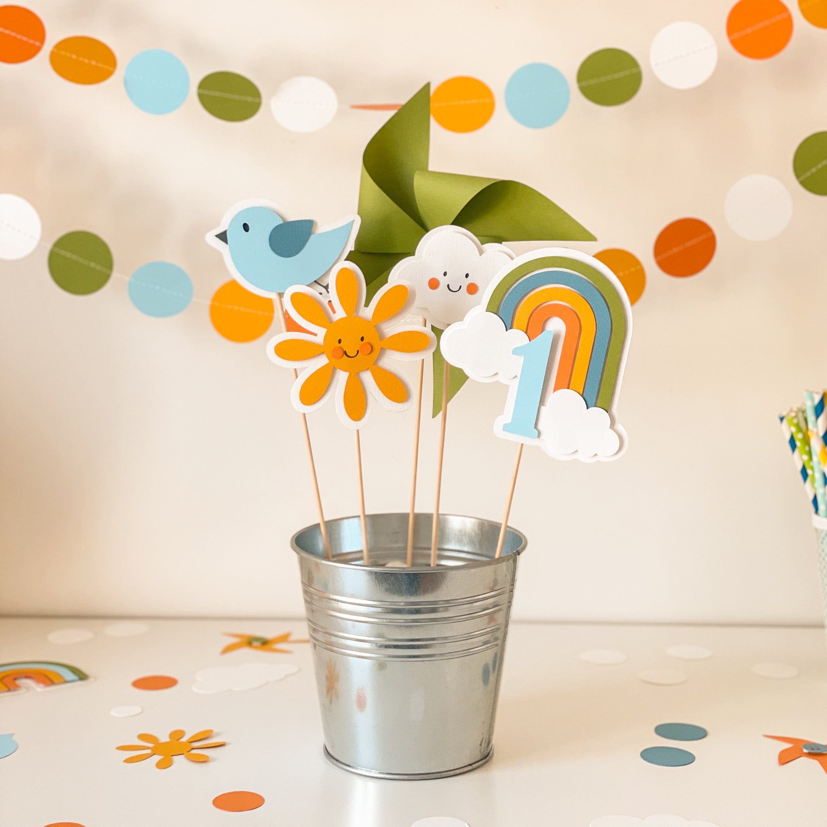 Decorative items including a number '1', rainbow, cloud, and sun in a metal bucket with colorful paper decorations in the background.