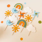 A set of colorful paper party decorations on wooden sticks, including two rainbows, two birds, two sunflowers, two clouds, and two windmill cutouts, arranged on a light beige background with scattered matching paper dots.