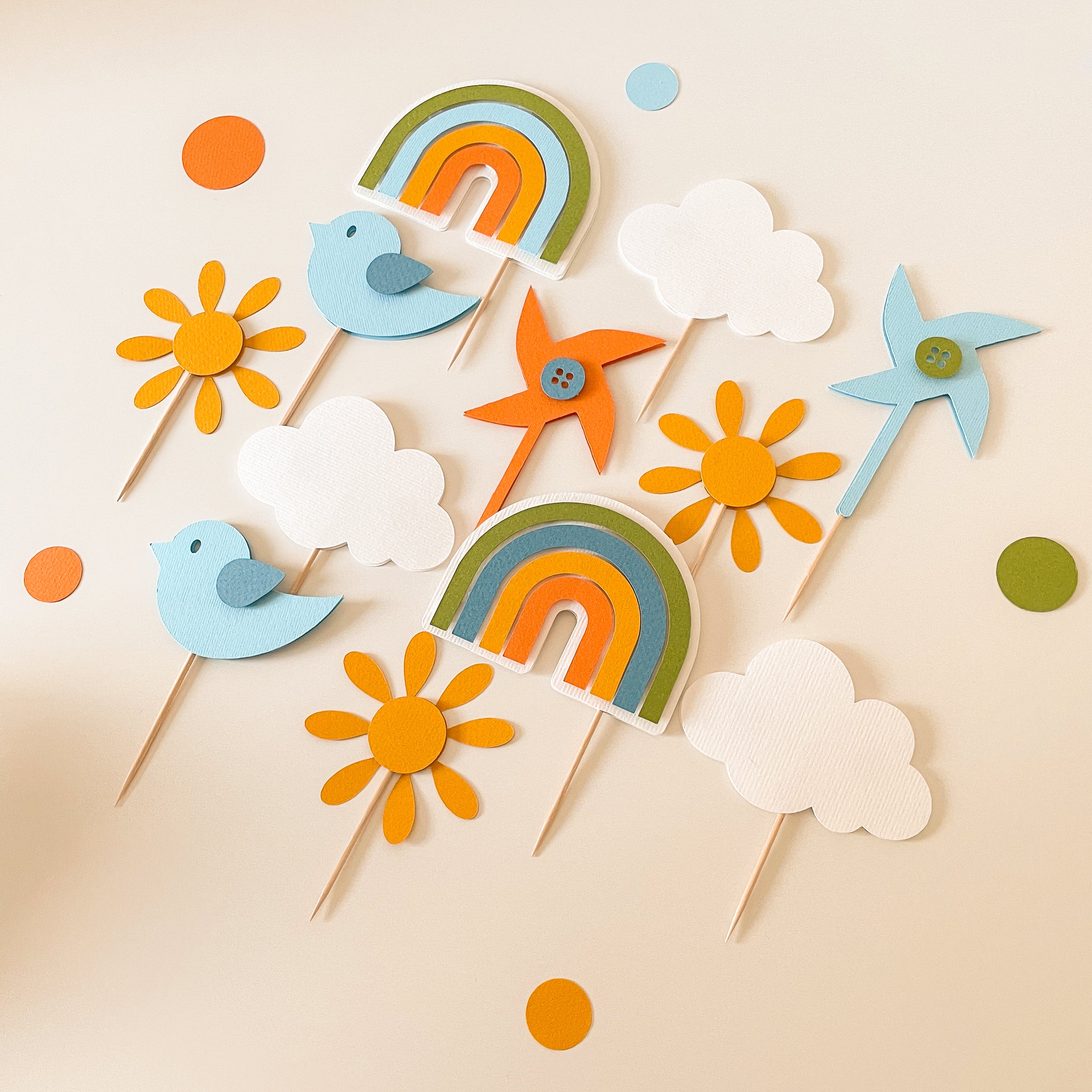 A set of colorful paper party decorations on wooden sticks, including two rainbows, two birds, two sunflowers, two clouds, and two windmill cutouts, arranged on a light beige background with scattered matching paper dots.