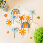 A set of colorful paper cutout decorations for a child's room, including rainbows, clouds, suns, birds, a windmill, and dots, arranged on a light beige surface.