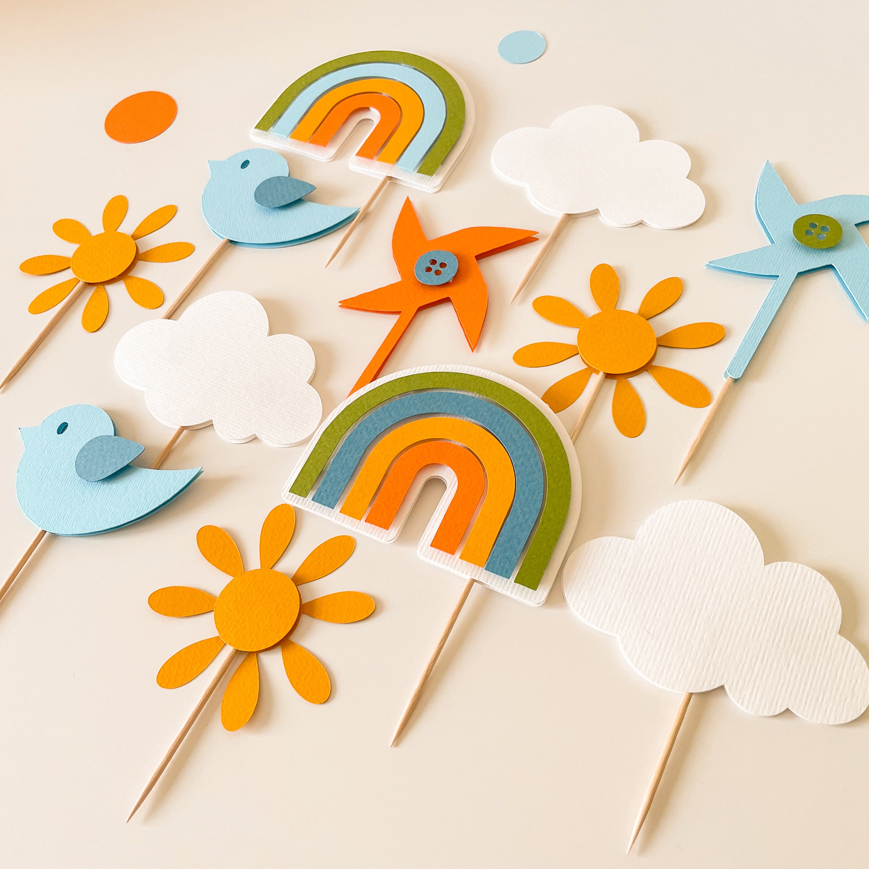 A set of colorful felt party cupcake toppers on wooden sticks, including rainbows, sunflowers, birds, clouds, a star, and a windmill, arranged on a light beige background.