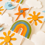 A flat-lay arrangement of colorful felt party decorations on wooden sticks, including a rainbow, sunflowers, clouds, a bird, and a pinwheel, on a light cream background.
