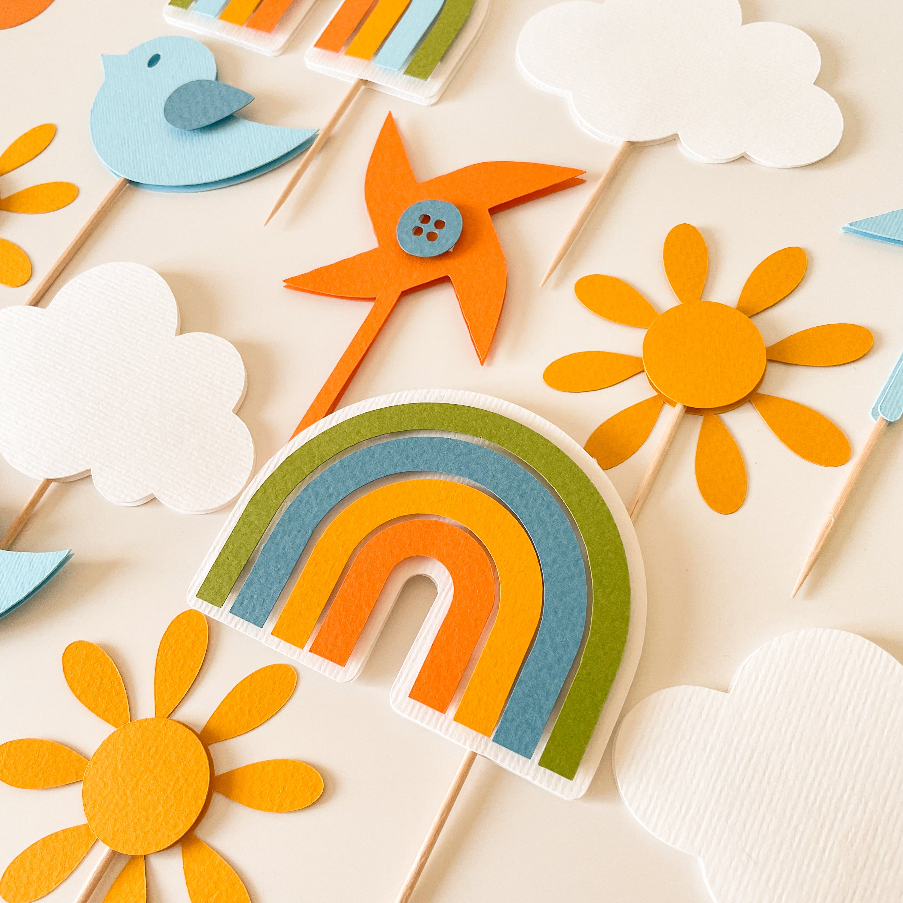 A flat-lay arrangement of colorful felt party decorations on wooden sticks, including a rainbow, sunflowers, clouds, a bird, and a pinwheel, on a light cream background.