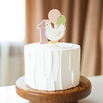 Small white cake with a chicken and balloon topper on a wooden stand.