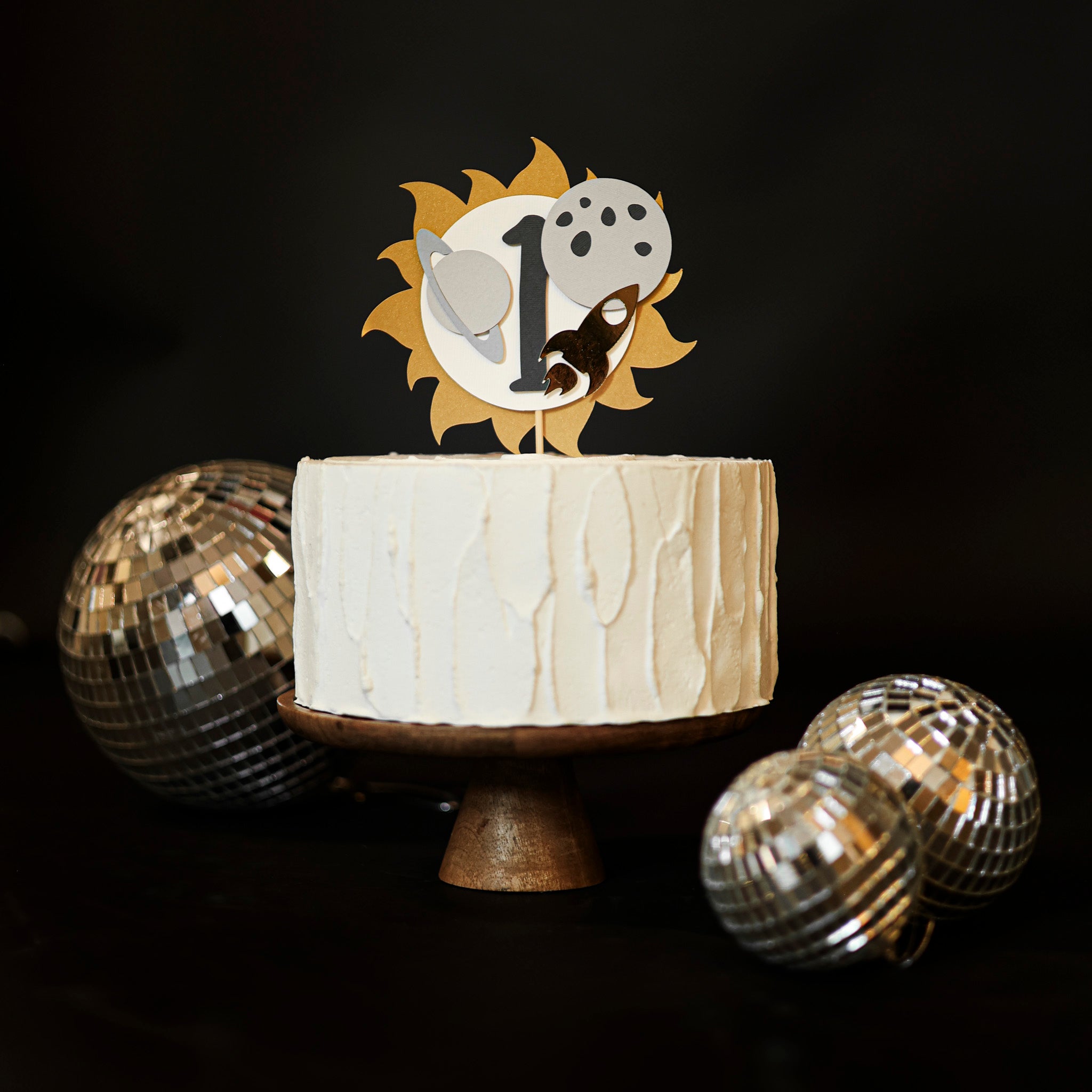 First Birthday cake with a decorative Space topper featuring a number '1', a golden sun, grey planet, moon, and gold rocket ship