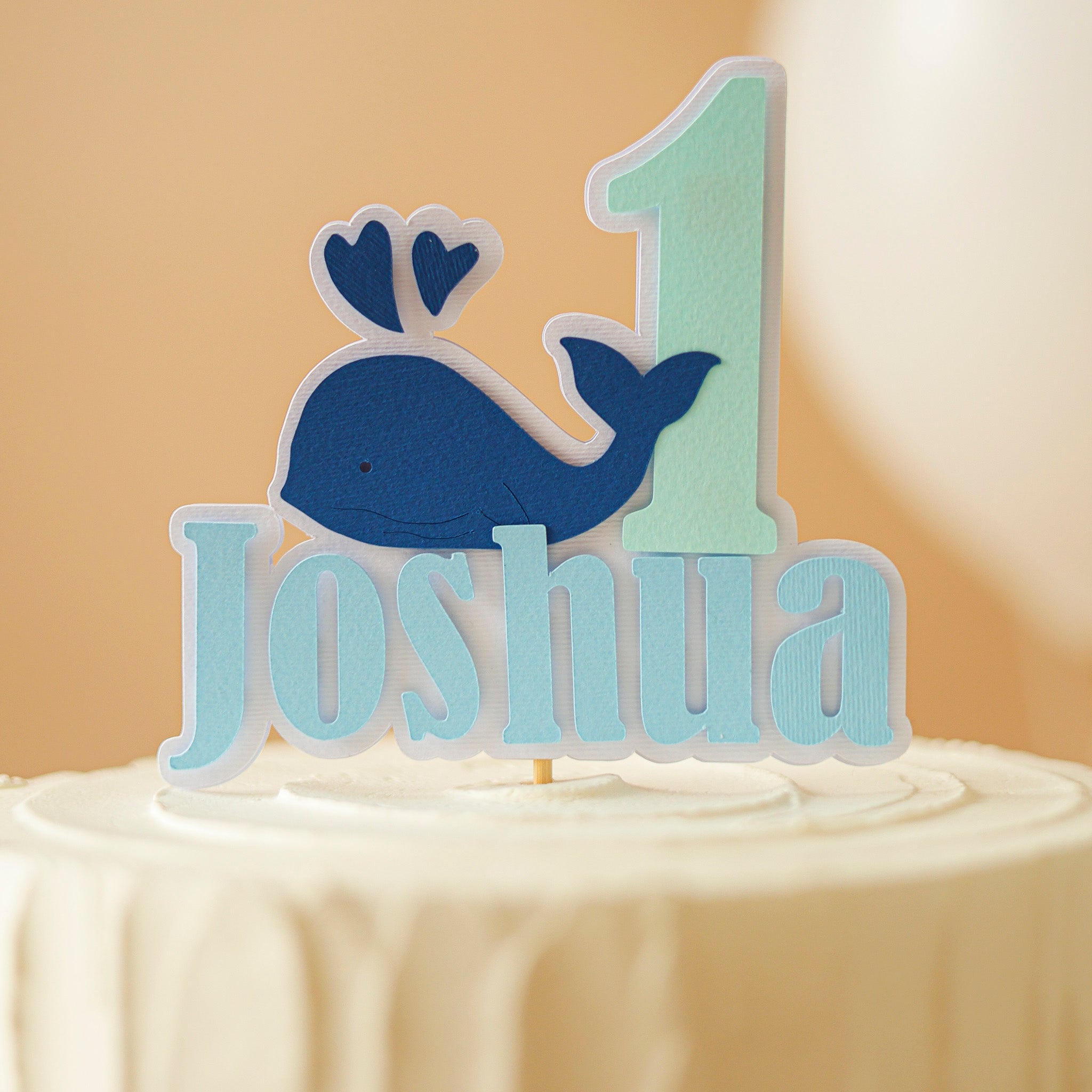 Cake topper with whale, number 1, and name 'Joshua' on a beige background