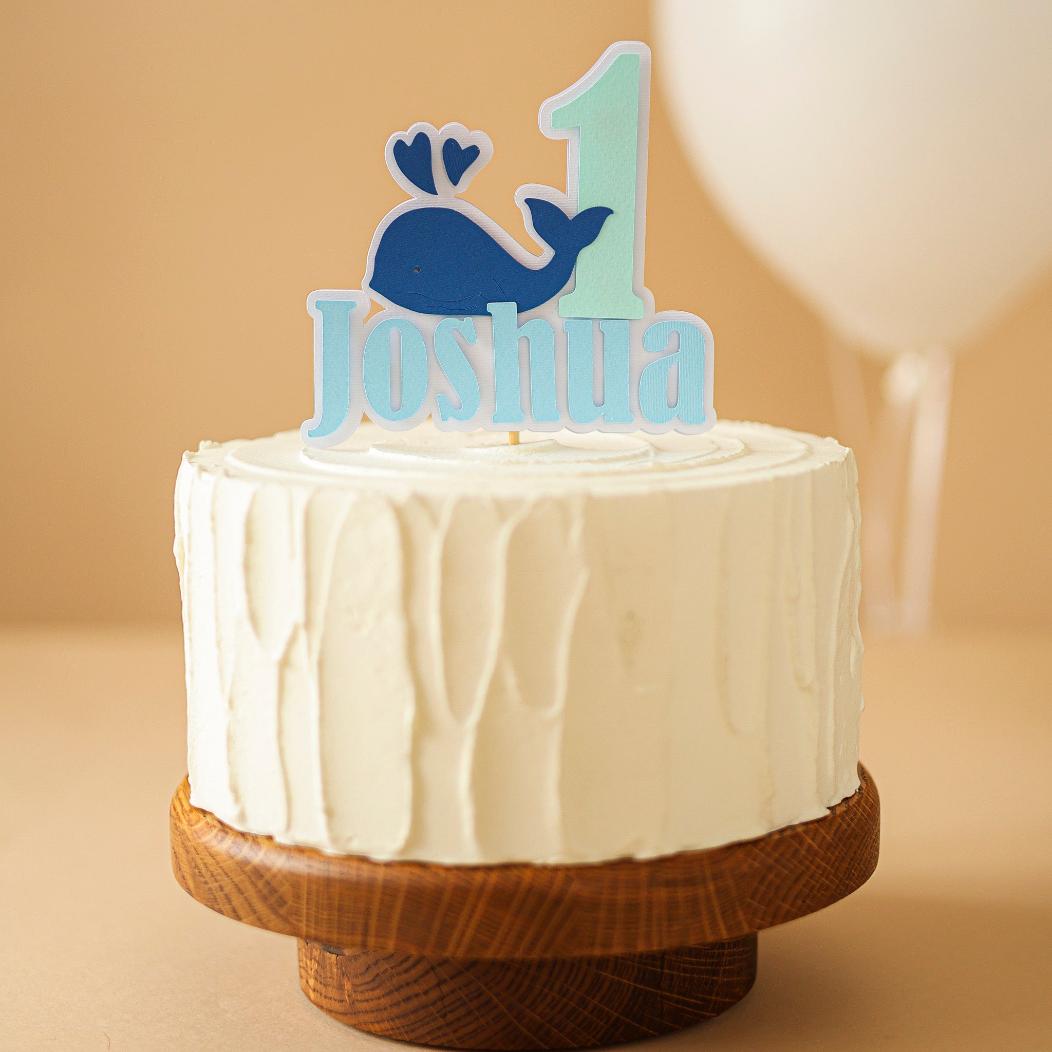 White cake with a whale-themed '1 Joshua' topper on a wooden stand 